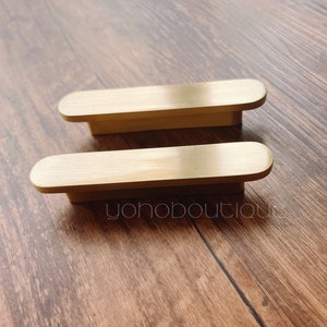 Könnte beinhalten: Zwei goldfarbene Schrankgriffe mit rechteckigem Design und abgerundeten Enden. Die Griffe sind auf einer Holzoberfläche platziert. Der Text "yohoboutique" ist sichtbar.