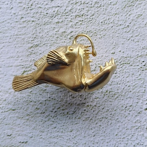 May include: A gold-colored metal pendant shaped like a deep-sea anglerfish with a large mouth and a lure.