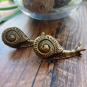 May include: Two antique brass snail ornaments with detailed shell patterns. The snails are on a wooden surface, with a glass bowl and greenery in the background. The ornaments are a decorative item.