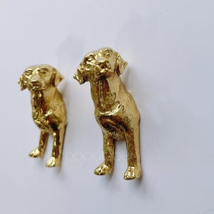 May include: Two gold-colored metal sculptures of Labrador Retriever dogs. The sculptures are detailed and realistic, with the dogs standing in a natural pose.