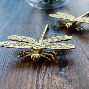 Puede incluir: Dos figuritas de libélula de metal dorado con alas y patas detalladas. Las libélulas están sobre una superficie de madera.