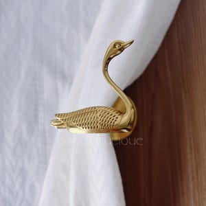 May include: A gold-coloured swan curtain tieback. The swan has a long, curved neck and detailed feathering on its body. The tieback is made of a shiny, metallic material and is designed to hold curtains in place.