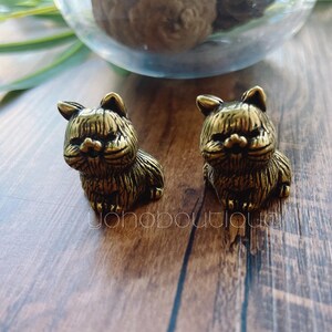 May include: Two small, bronze-colored cat figurines sit on a wooden surface. The cats are detailed with textured fur and have a slightly stern expression. A glass bowl with decorative items is in the background.