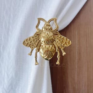 以下が含まれることがあります： 金色の金属製のハチのカーテンタッセル。ハチは詳細な羽と体のセグメントを持ち、白いカーテンに取り付けられています。カーテンタッセルは、家の装飾用の装飾要素です。