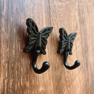 May include: Two black butterfly-shaped hooks mounted on a wooden surface. The hooks have a decorative design and a simple, curved shape.