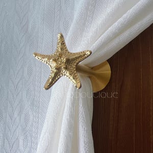 Peut inclure: Un embrasse-rideau en forme d'étoile de mer dorée. L'étoile de mer est texturée et montée sur une base circulaire dorée. L'embrasse-rideau maintient un rideau blanc. L'arrière-plan comprend une porte en bois et un mur blanc texturé.