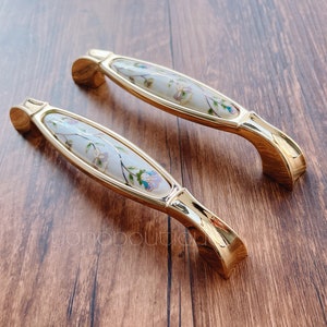 Könnte beinhalten: Zwei goldene Schrankknöpfe mit einer weißen Keramikbasis und einem Blumendesign.