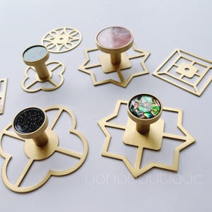 May include: Five gold metal hooks with different geometric designs and decorative tops. The tops are made of various materials, including pink, black, and iridescent shell.