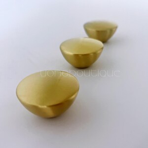 Könnte beinhalten: Drei goldfarbene, halbkugelförmige Schrankknöpfe, die diagonal auf einer weißen Oberfläche angeordnet sind. Die Knöpfe haben eine glatte, matte Oberfläche und eine abgerundete Form, was auf ein modernes Design hindeutet. Die Knöpfe sind wahrscheinlich für Möbel oder Dekorationszwecke bestimmt.