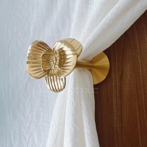 May include: A gold-coloured curtain tieback shaped like a flower, holding a white curtain. The tieback is mounted on a wooden surface. The flower has detailed petal designs.
