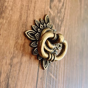 May include: A gold-toned decorative door or cabinet pull with a detailed floral design. The pull has a ring-shaped handle.