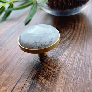 Könnte beinhalten: Ein Schrankknopf mit einer runden, hellgrauen Steinplatte mit weißem Marmormuster, eingebettet in eine goldfarbene Metallbasis. Der Knopf ist für Möbel konzipiert und hat ein klassisches, elegantes Aussehen.