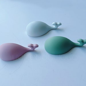Puede incluir: Tres figuritas de ballena de cerámica en diferentes colores: blanco, rosa y verde. Cada ballena tiene una forma simple y redondeada y una pequeña aleta de cola.