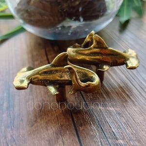 Könnte beinhalten: Zwei Schrankknöpfe aus Messing in Form von Hammerhaien. Die Knöpfe sind mit Flossen und Kiemen versehen und auf kurzen Pfosten montiert. Die Knöpfe haben eine warme Goldfarbe.