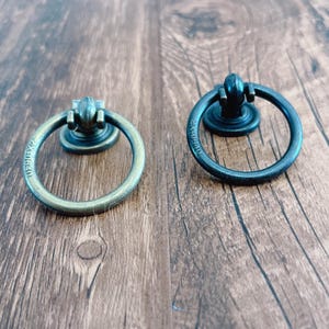 Op de afbeelding: Twee ringvormige kastgrepen, een in antiek messing en een in zwart. Beide hebben een decoratieve knop aan de bovenkant.