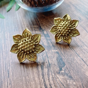 以下が含まれることがあります： 真鍮色の花の形をしたキャビネットノブが2つ。各ノブは、花びらとテクスチャ加工された中心部を備えた詳細な花の模様が特徴です。ノブは木製の表面に表示され、装飾的な魅力を示しています。