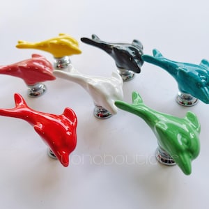 May include: Set of 7 ceramic dolphin door knobs in various colors, including red, yellow, green, blue, white, and gray. The knobs are shaped like dolphins with a smooth, glossy finish.