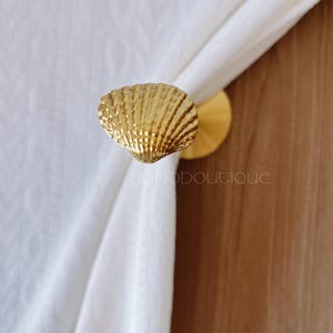 May include: A gold-colored seashell curtain tieback. The tieback is made of metal and has a textured surface. The curtain tieback is holding a white curtain. The text "uonoboutique" is visible.
