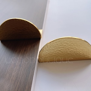 Puede incluir: Dos tiradores de armario de media luna de color dorado con textura martillada.