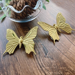 May include: Two gold metal butterfly shaped decorative accents. The butterflies are on a wooden surface.