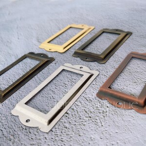 May include: Five rectangular label holders in various metallic finishes: gold, bronze, copper, and silver. Each frame has a decorative top edge and two small holes for mounting. These vintage-style label holders are ideal for drawers or cabinets.