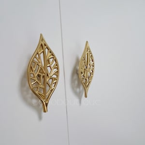 May include: Two gold leaf-shaped cabinet knobs with a delicate, intricate design. The knobs are mounted on a white surface.