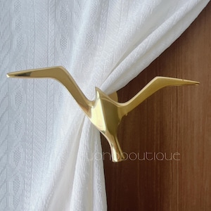 May include: A gold-coloured metal curtain tieback shaped like a bird, holding white fabric. The tieback is attached to a wooden surface. The word "uonboutique" is visible on the wooden surface.