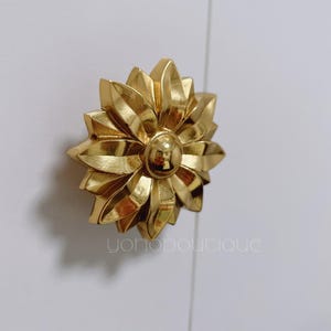 Könnte beinhalten: Vergoldeter, blumenförmiger Schrank-Knopf aus Metall mit strukturierter Oberfläche.