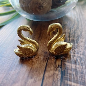 以下が含まれることがあります： 2つの金色の白鳥の置物。白鳥は互いに向き合い、首を曲げ、翼をわずかに広げています。置物は木製の表面にあり、背景はぼやけています。