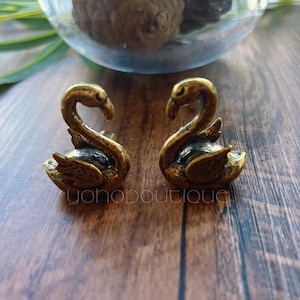 Op de afbeelding: Twee kleine, antiek goudkleurige zwaanbeeldjes. De zwanen kijken elkaar aan, met gebogen halzen en licht gespreide vleugels. De beeldjes staan op een houten oppervlak. De tekst "yohoboutique" is zichtbaar.