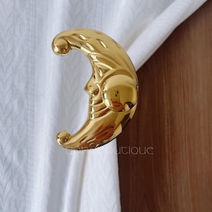 Könnte beinhalten: Ein goldfarbener Gardinenhalter in Form eines Halbmonds. Der Halter hat ein detailliertes Gesichtsdesign und hält einen weißen Vorhang. Der Text "yohana boutique" ist auf dem Vorhang zu sehen.