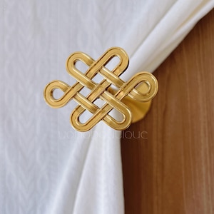 Peut inclure: Un embrasse-rideau doré avec un motif de nœud entrelacé complexe. L'embrasse est fixée sur une base circulaire et maintient un rideau blanc. L'arrière-plan comprend une surface en bois.