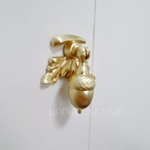 Op de afbeelding: Een gouden deurklopper in de vorm van een eikel met een bladontwerp. De deurklopper is bevestigd aan een witte deur.