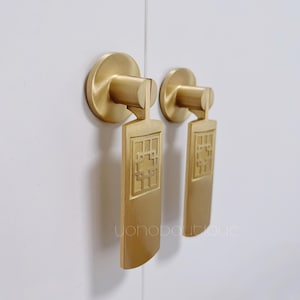 May include: Two gold cabinet pulls with a rectangular design featuring a geometric pattern. The pulls are mounted on a white surface.