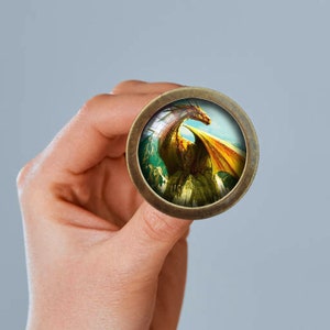 Op de afbeelding: Een ronde, bronskleurige knop met een gedetailleerde afbeelding van een draak. De draak heeft oranje en gele vleugels, met een groene en blauwe achtergrond. De knop wordt in een hand gehouden.