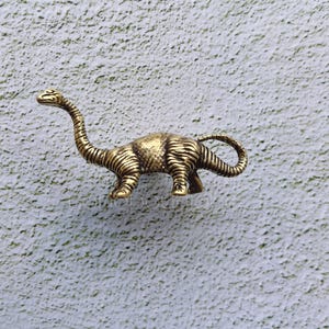 Puede incluir: Una figurita de dinosaurio de color dorado con cuello y cola largos. El dinosaurio está de pie sobre una superficie blanca texturizada.