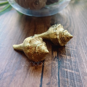 May include: Two gold-colored seashell ornaments are displayed on a wooden surface. The seashells have detailed ridges and a spiral shape. The image is well-lit, highlighting the metallic sheen of the ornaments. The shop's name, "yonoboutique", is visible in the bottom right corner.