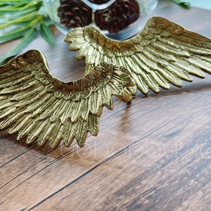 May include: A pair of gold-colored metal angel wings. The wings are detailed with intricate feather patterns and are arranged in a crossed position.