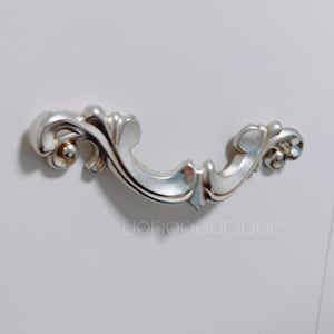 Puede incluir: Un tirador de cajón de plata adornado con un diseño decorativo en espiral. El tirador está montado en una superficie blanca.