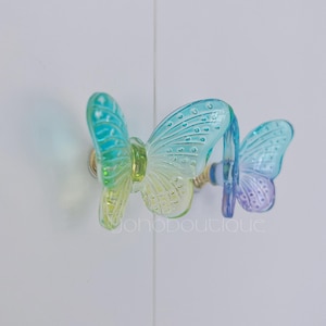 Op de afbeelding: Twee vlindervormige kastknoppen. De knoppen zijn gemaakt van doorschijnend glas met een kleurovergang van blauw, groen en geel. De knoppen zijn gemonteerd op goudkleurige hardware. Het algemene thema is decoratieve woondecoratie.