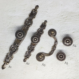 May include: Five antique-style cabinet hardware pulls in a brass finish. The pulls feature ornate designs and include a long pull, a shorter pull, a D-shaped pull, and two round knobs.