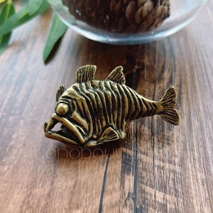 May include: A gold-coloured metal figurine of a fish with a detailed, textured surface. The fish has a large, open mouth and a long, slender body.