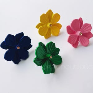 May include: Four flower-shaped metal embellishments in different colours: blue, yellow, pink, and green. Each flower has a small rhinestone in the centre.