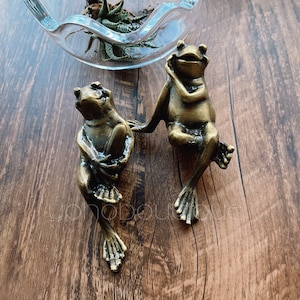 Może przedstawiać: Dwie figurki żab z brązu, jedna z ręką na głowie, druga z ręką na brodzie. Żaby znajdują się na drewnianej powierzchni, z szklaną miską i zielenią w tle. Żaby są szczegółowe z teksturowaną skórą.