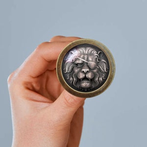 Peut inclure: Une épingle ronde de couleur laiton avec une image détaillée en trois dimensions d'un visage de lion. Le lion a un cache-œil noir sur son œil droit.