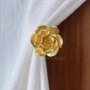 May include: A gold-coloured, rose-shaped curtain tieback. The tieback is made of metal and has detailed petals. The text "yonoboutique" is visible at the bottom of the image. The tieback is holding a white curtain.