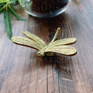 Puede incluir: Una figurita de libélula de color dorado con alas detalladas. La figurita está sobre una superficie de madera.