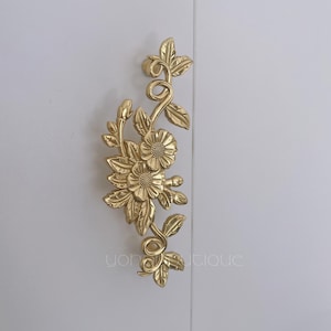 Puede incluir: Un tirador de armario floral dorado con detalles intrincados. El tirador presenta un grupo de flores, hojas y zarcillos. El herraje está montado sobre una superficie blanca. El texto "yon boutique" es visible.