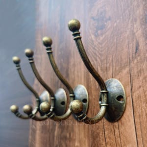 May include: A set of five antique-style wall hooks in a warm brass finish. Each hook features a curved design with a decorative ball at the end and a round mounting plate. The hooks are mounted on a wooden surface. The text "yohoboutique" is visible.
