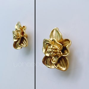 Pode incluir: Dois botões de armário em forma de flor dourados. Os botões têm um design detalhado e texturizado.